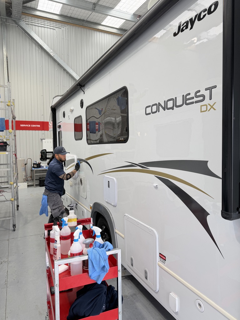 Detailer hand-polishing the sidewall of a Jayco Conquest DX motorhome with a trolley of Koch-Chemie products in the Auckland workshop